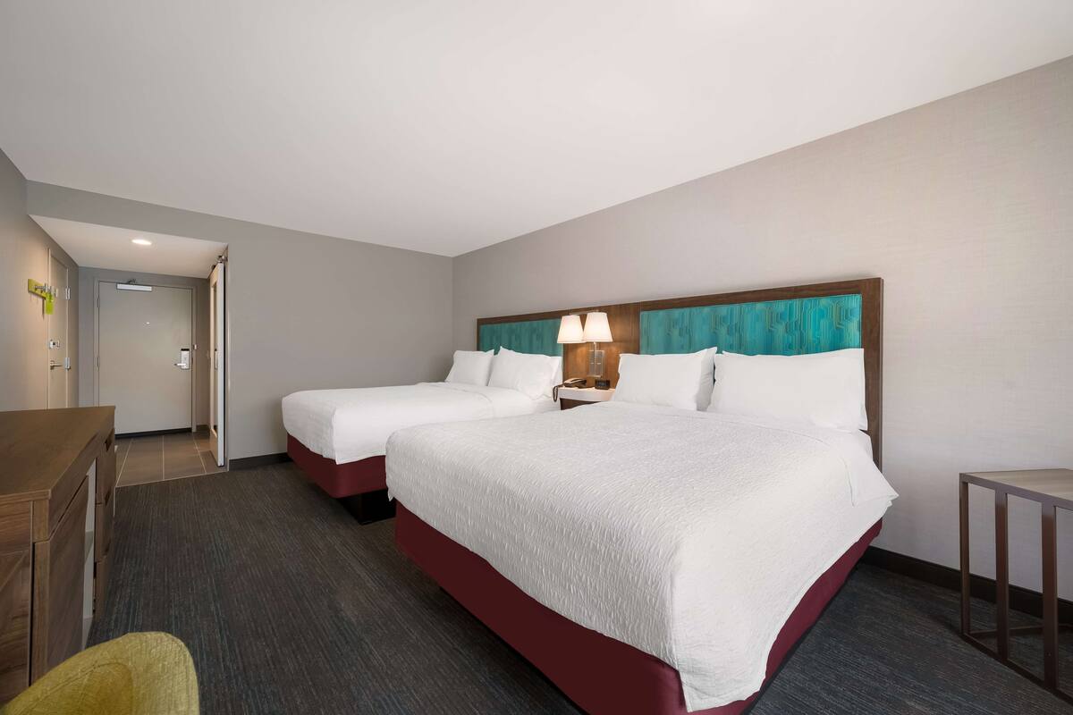 A hotel room with two beds, a desk, a chair, and a nightstand with a lamp.