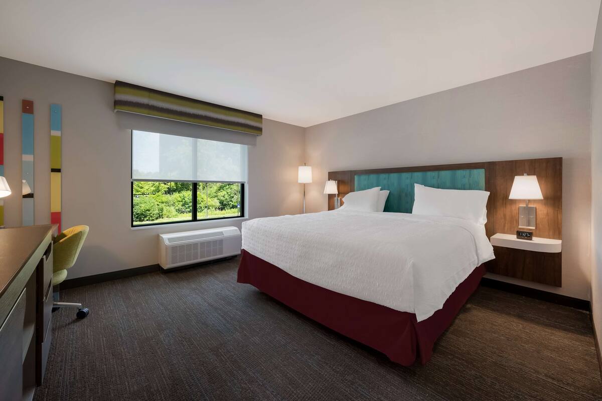 A hotel room with a large bed, two bedside lamps, a desk with a chair, a window with a view of greenery, and a wall with abstract art.