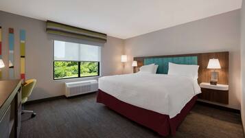 A hotel room with a large bed, two bedside lamps, a desk with a chair, a window with a view of greenery, and a wall with abstract art.