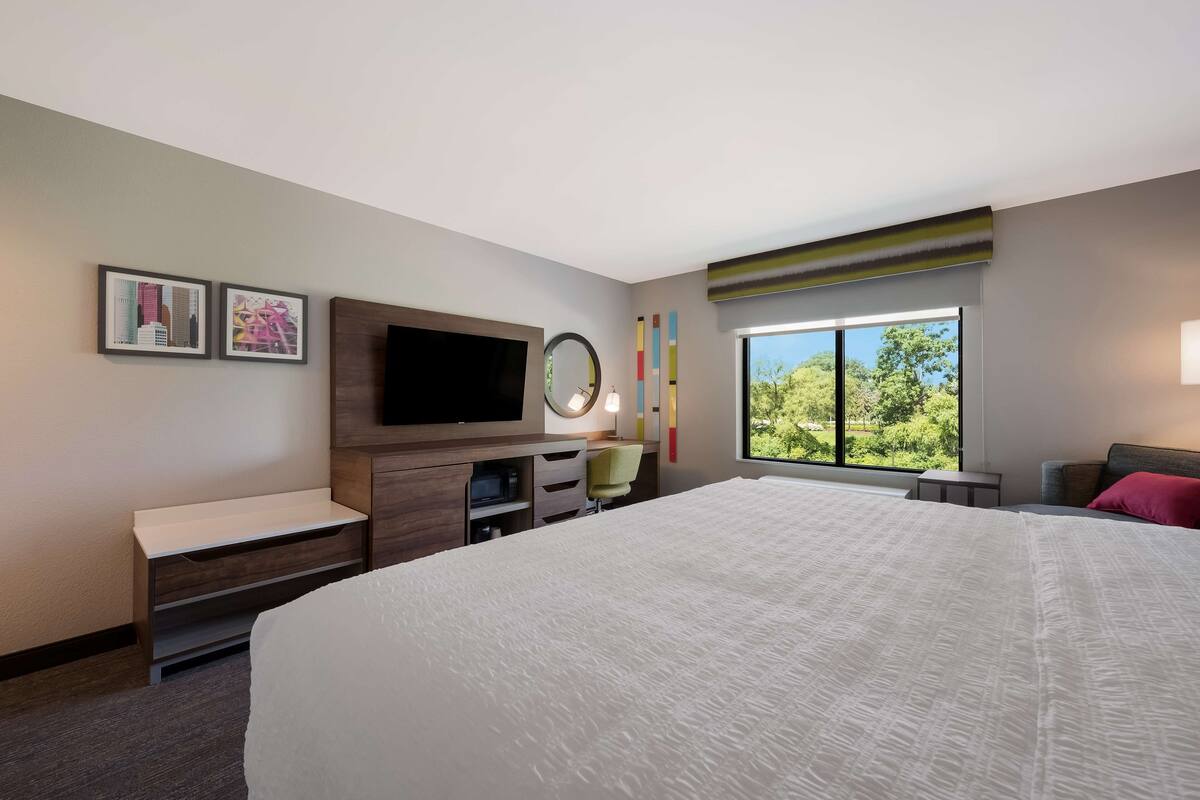 A hotel room with a large bed, a TV, a desk with a chair, a small table, and a window with a view of greenery.
