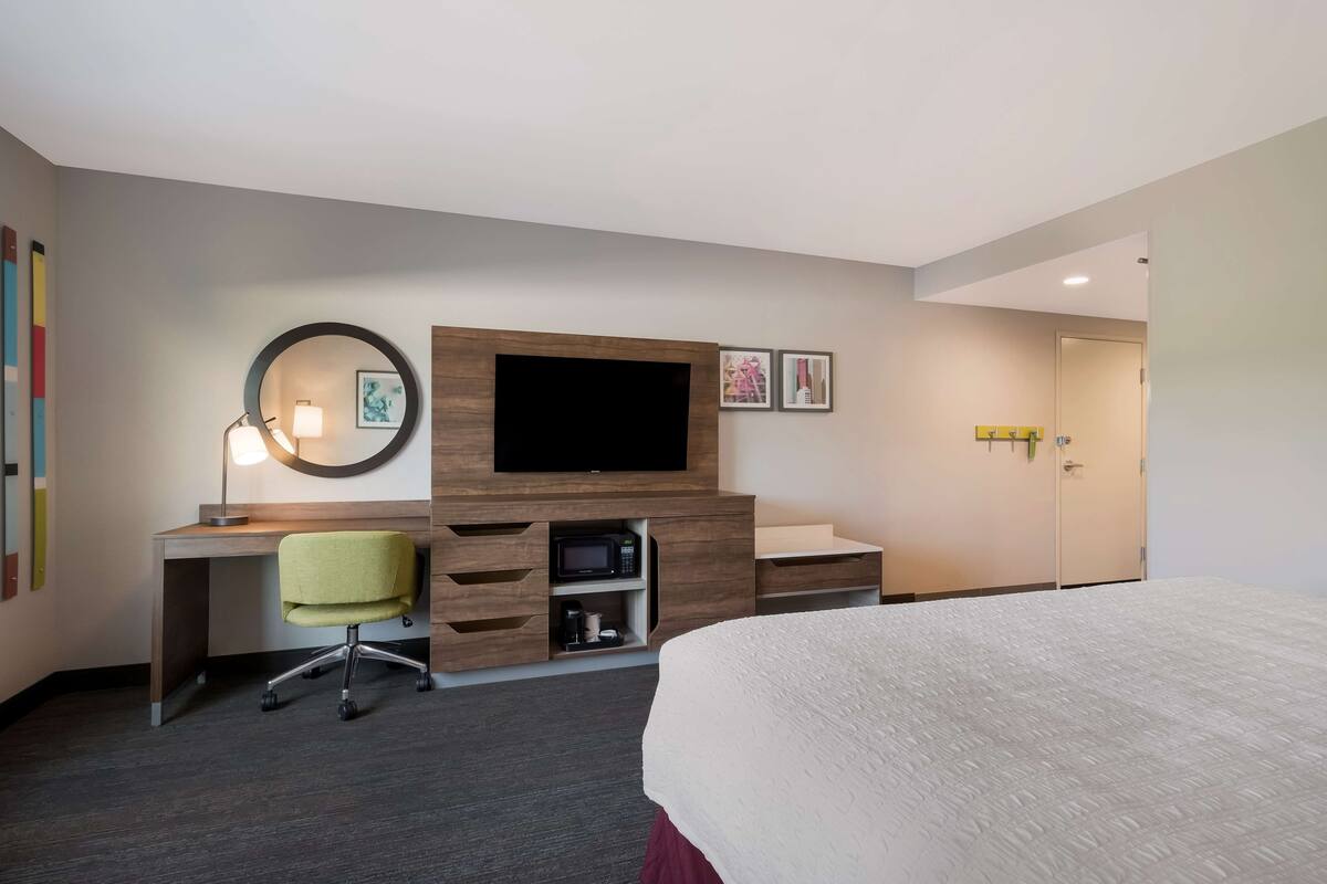 A hotel room with a bed, a desk with a chair, a TV, a microwave, and a door.