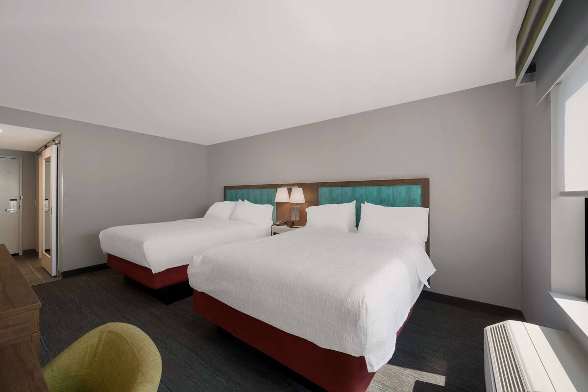 A hotel room with two beds, a desk, a chair, and a window.