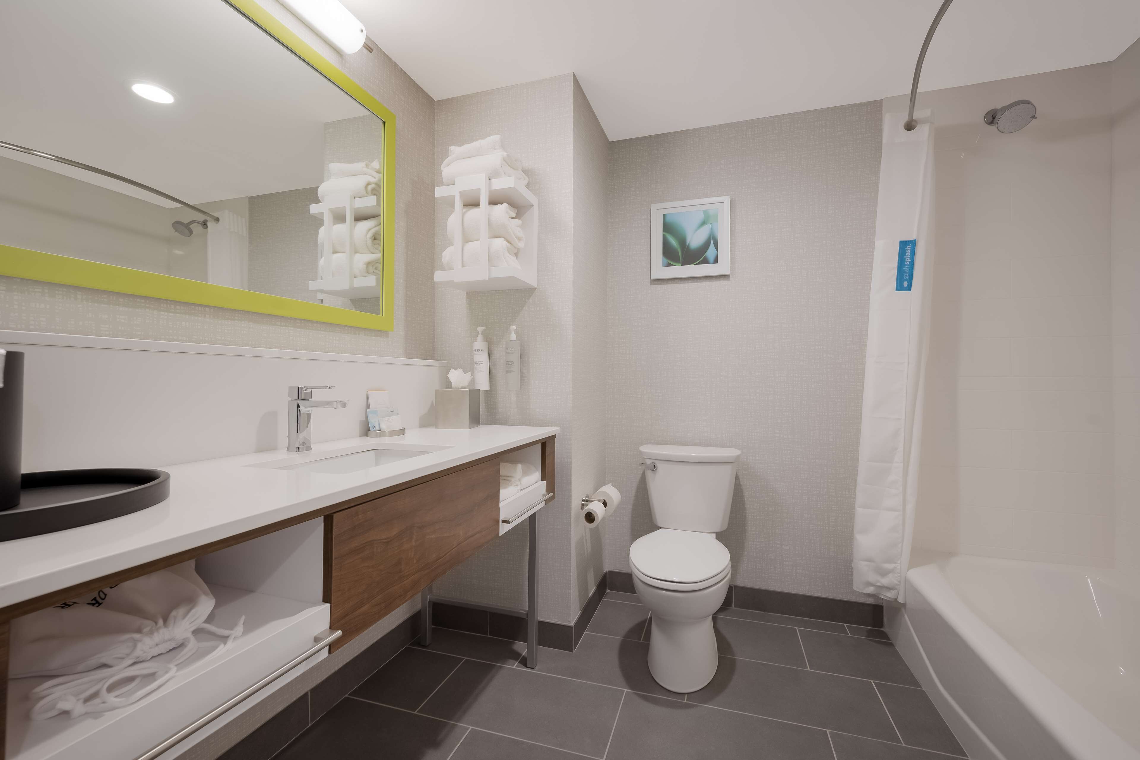 A modern bathroom with a white sink, toilet, and bathtub. There is a mirror with a yellow frame, a towel rack, and a framed picture on the wall.