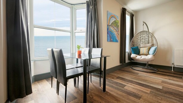 Een moderne kamer met een glazen tafel, zwarte stoelen, een hangstoel en uitzicht op zee.