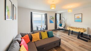 Een moderne woonkamer met een bank, kleurrijke kussens, een tafelvoetbaltafel en uitzicht op zee door grote ramen.