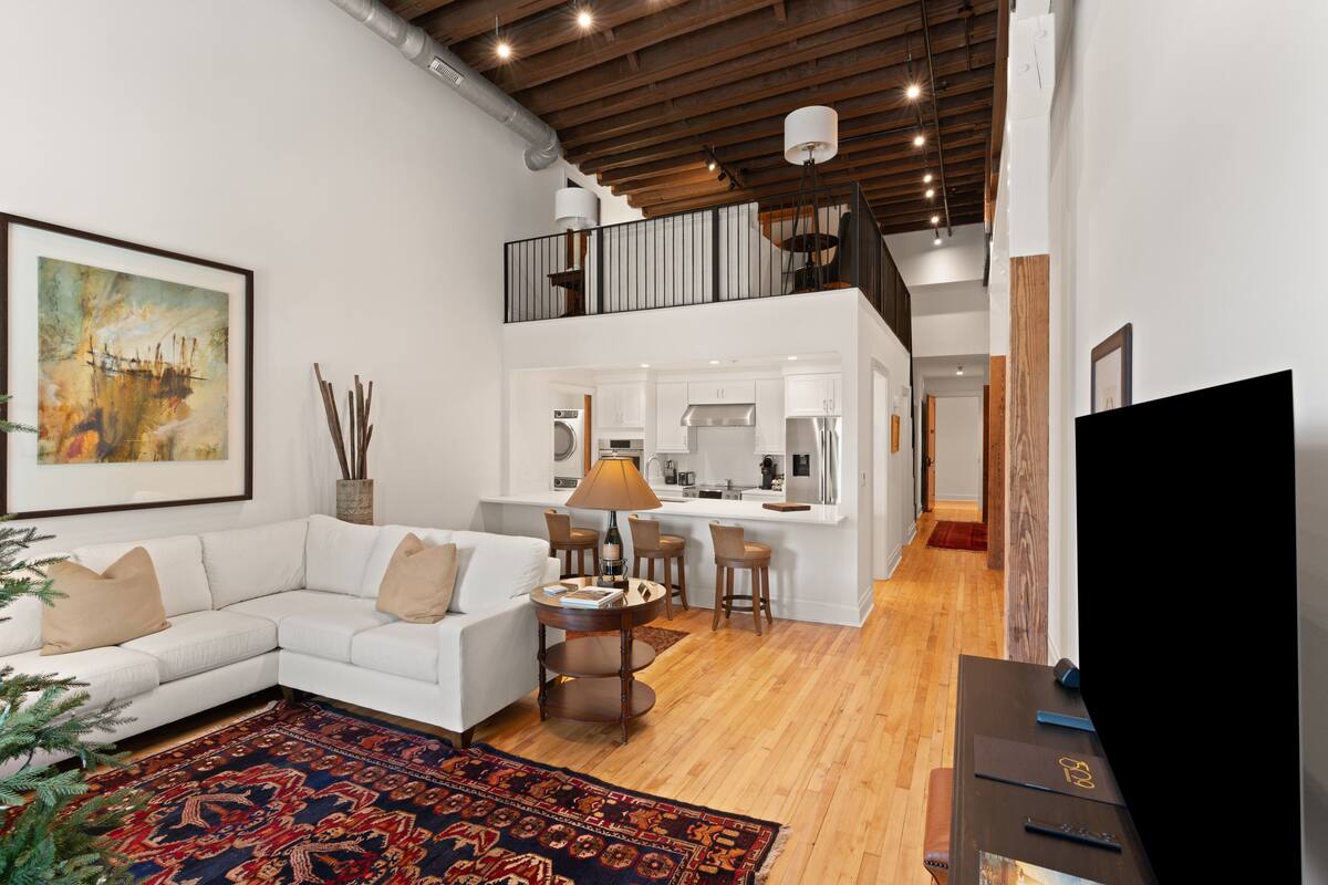 A modern living room with a white sofa, a coffee table, and a large rug. There is a kitchen area with a dining table and chairs. A TV is visible on the right side.