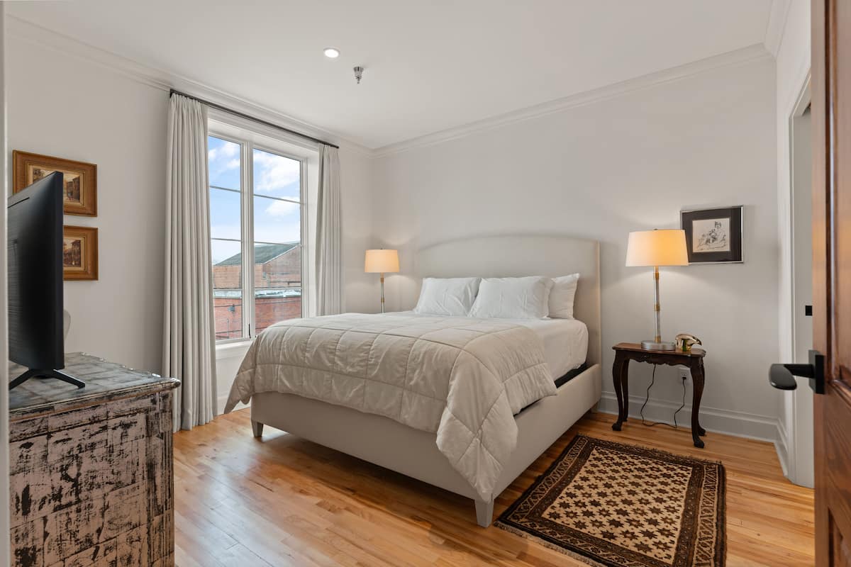 A bedroom with a bed, two bedside lamps, a television on a stand, and a window with curtains.