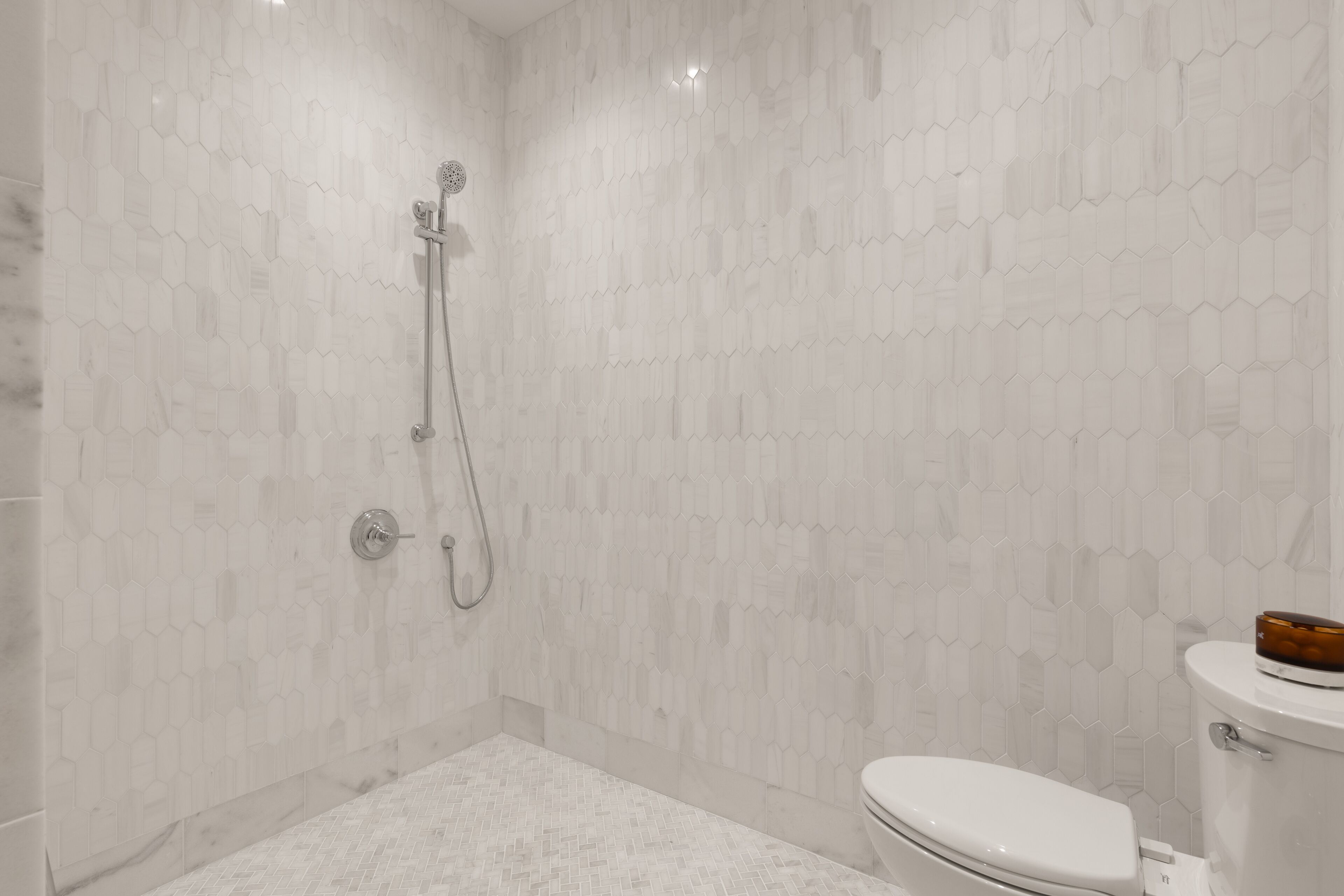 A modern bathroom with a hexagonal tile pattern, a shower with a handheld showerhead, and a toilet with a brown bowl.