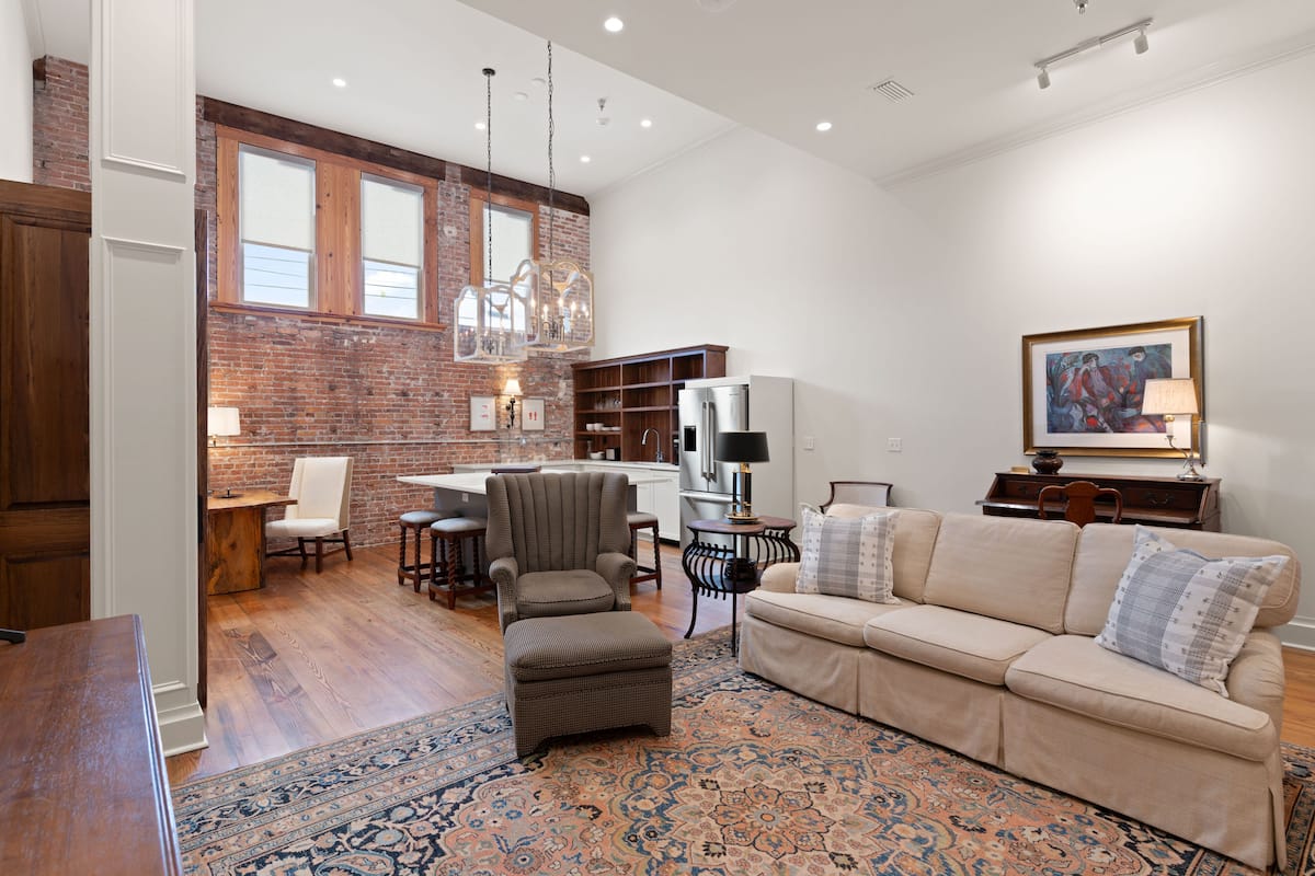 A spacious living area with a large sofa, ottoman, and a patterned rug. A kitchen with a brick wall and modern appliances is visible in the background.