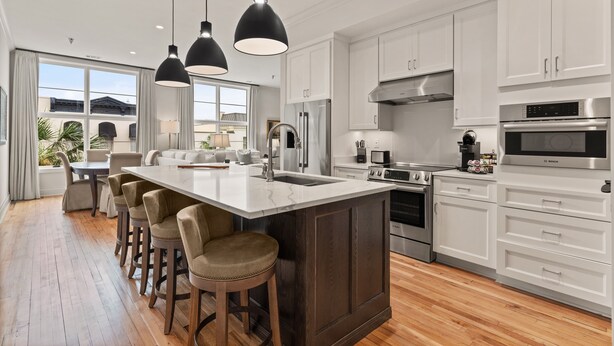 A modern kitchen with white cabinets, a large island with stools, stainless steel appliances, and pendant lighting. - The Kress Brunswick (Brunswick)