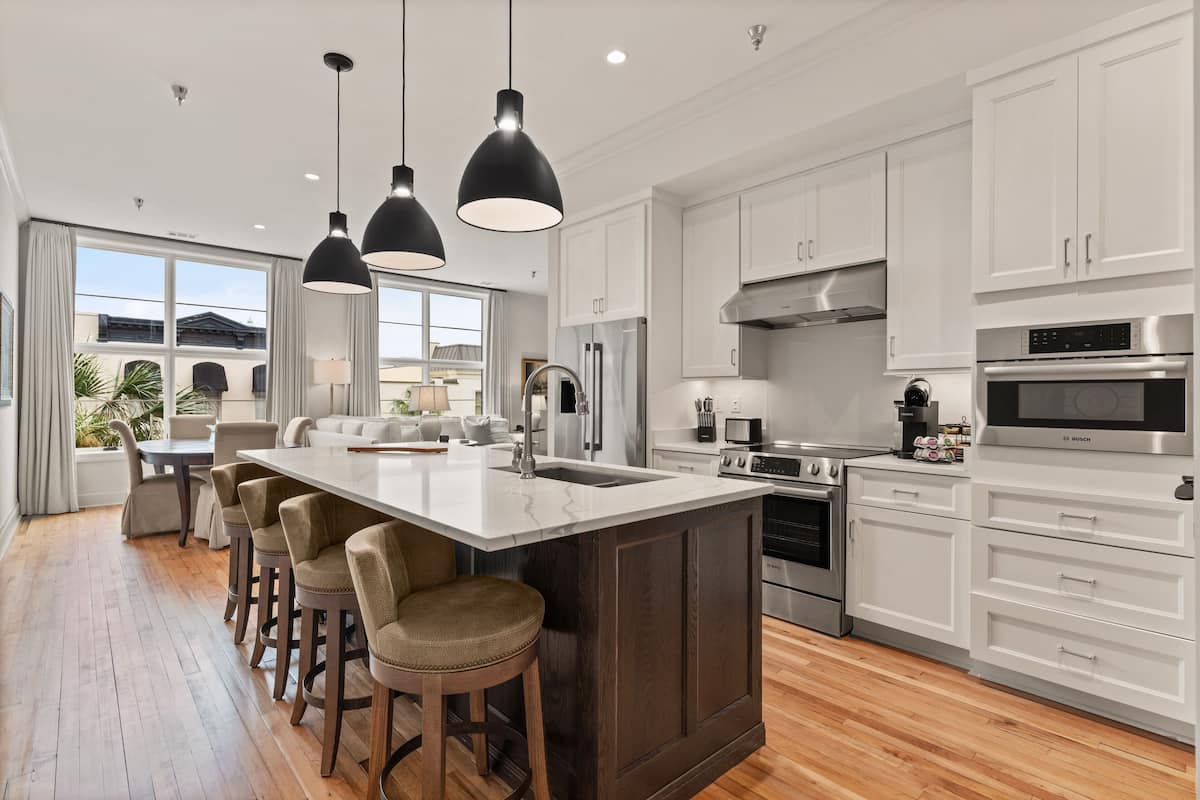 A modern kitchen with white cabinets, a large island with stools, stainless steel appliances, and pendant lighting.