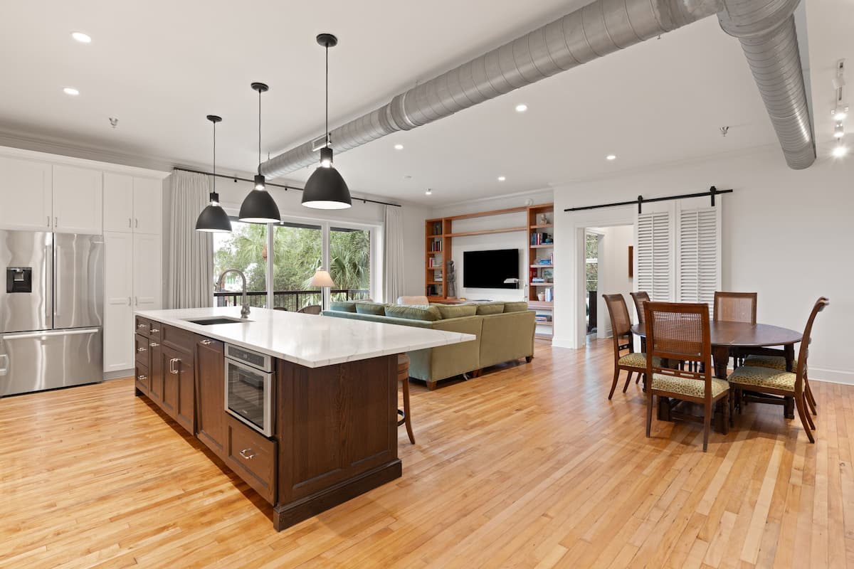 A spacious kitchen with a large island, wooden dining table, and chairs, a living area with a sofa and TV, and a view of greenery outside.