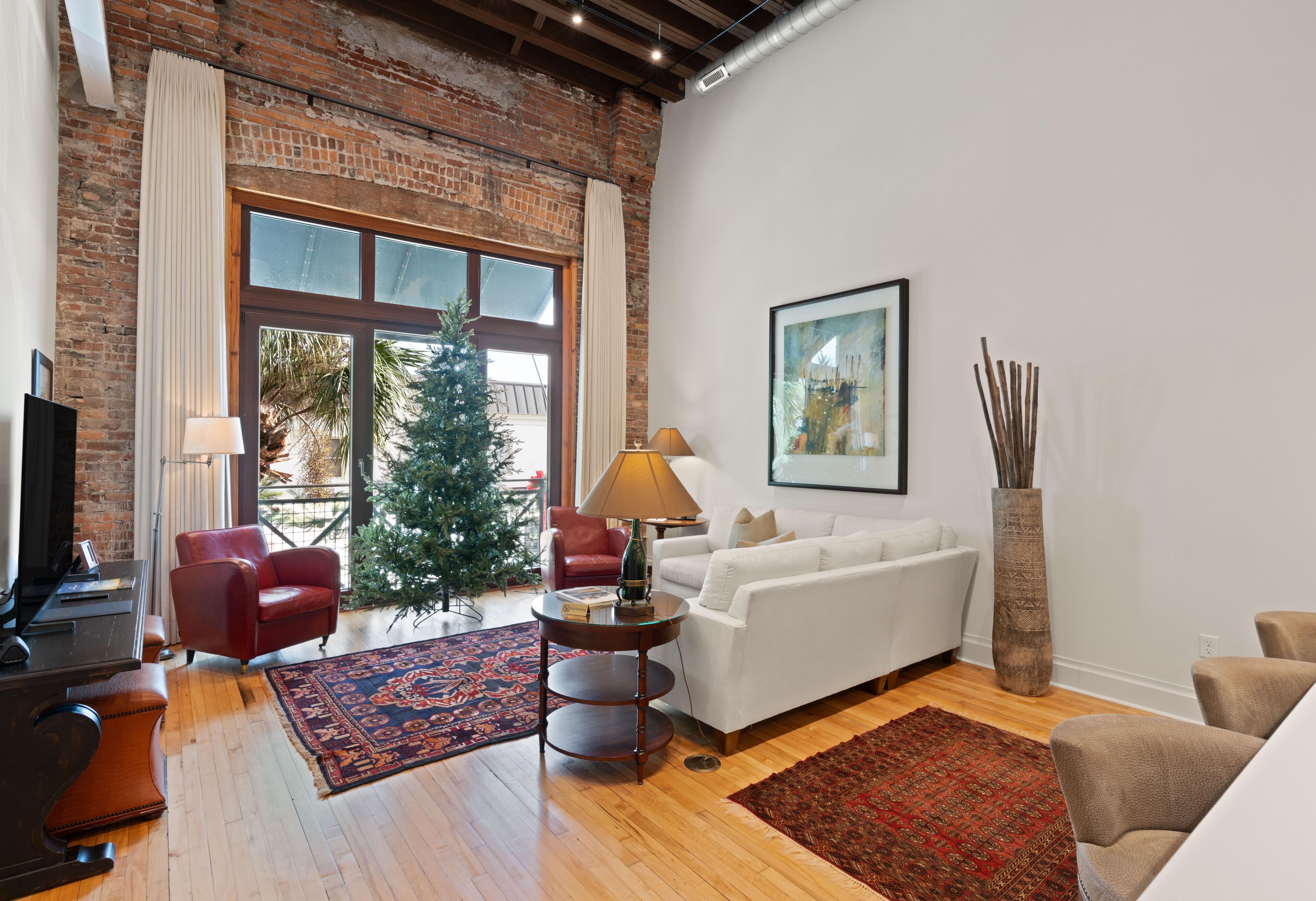 A living room with a brick wall, a white sofa, a red chair, a coffee table, a lamp, a tall vase, and a Christmas tree.