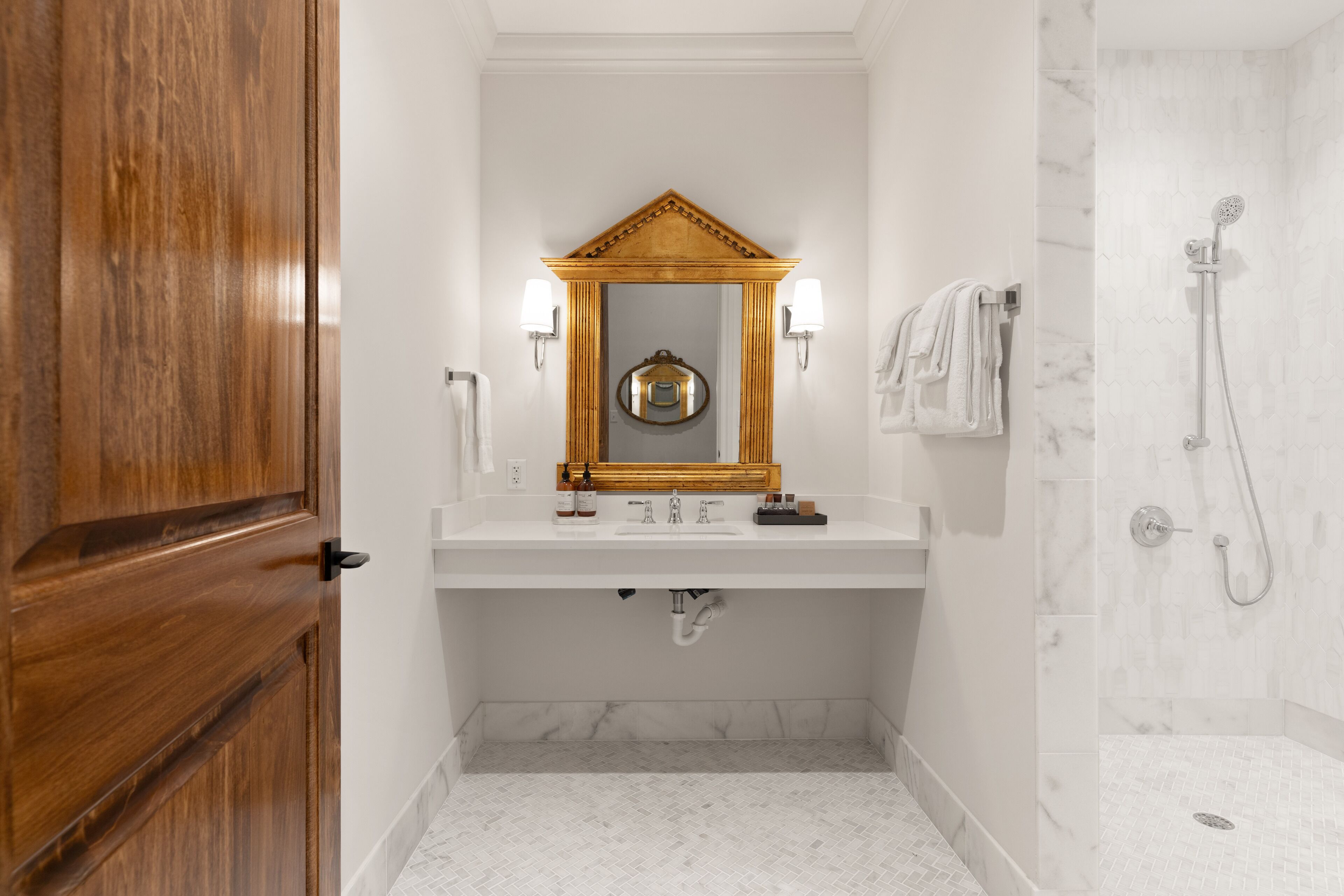 A bathroom with a wooden door, a white sink with a mirror above it, two wall-mounted lights, a towel rack with towels, and a shower area with a marble wall.
