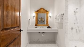 A bathroom with a wooden door, a white sink with a mirror above it, two wall-mounted lights, a towel rack with towels, and a shower area with a marble wall.