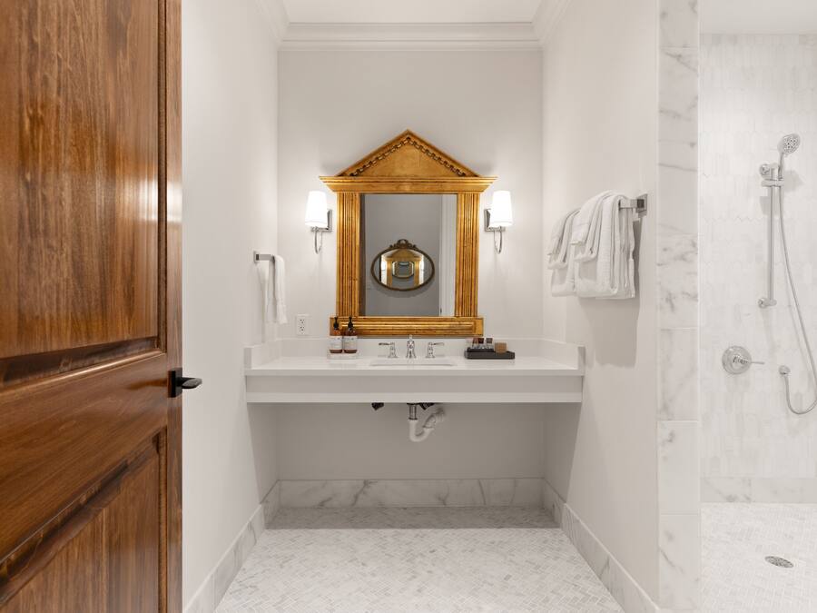 A bathroom with a wooden door, a white sink with a mirror above it, two wall-mounted lights, a towel rack with towels, and a shower area with a marble wall.