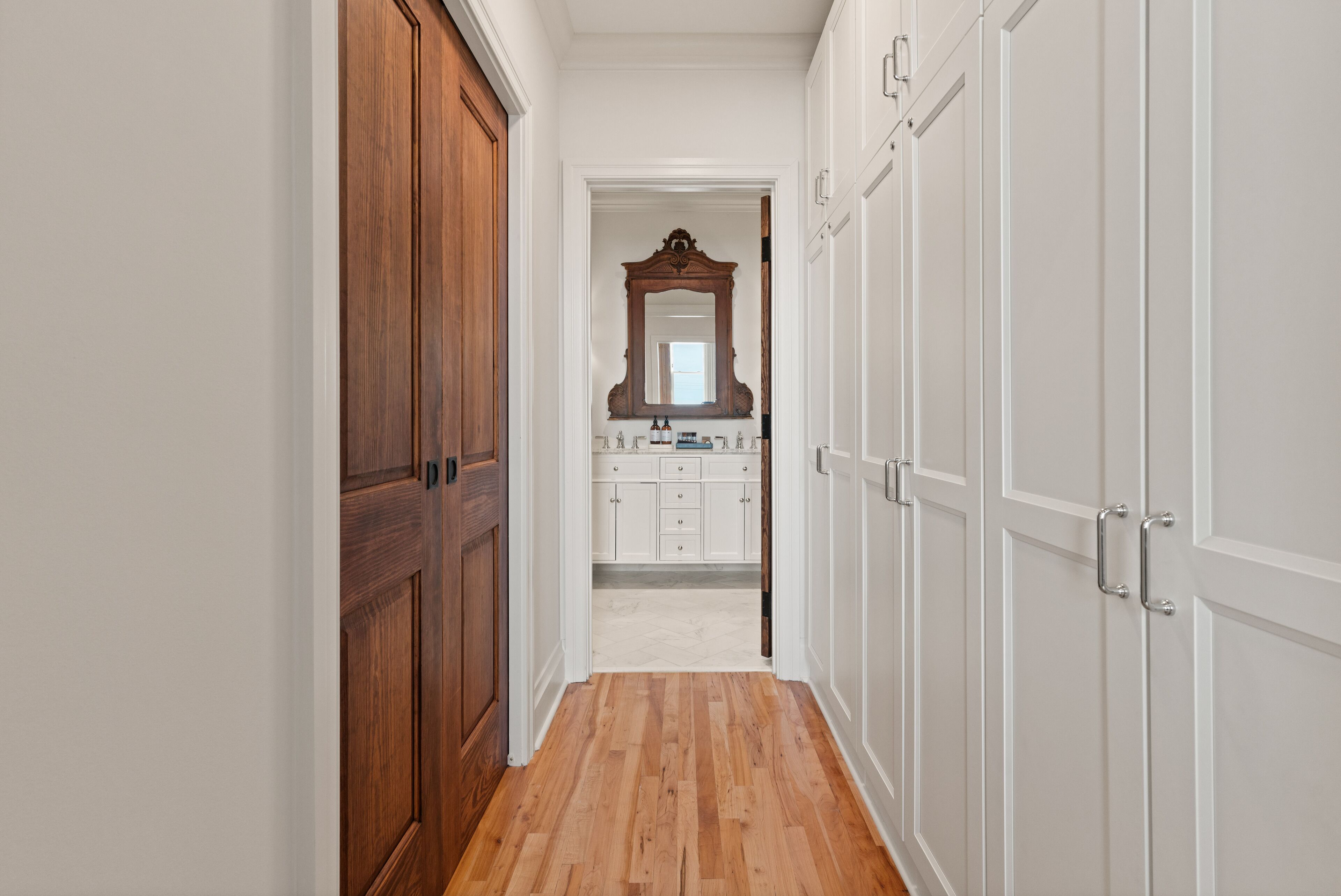 A narrow hallway with wooden doors on the left and white doors on the right, leading to a bathroom with a large mirror and cabinets.