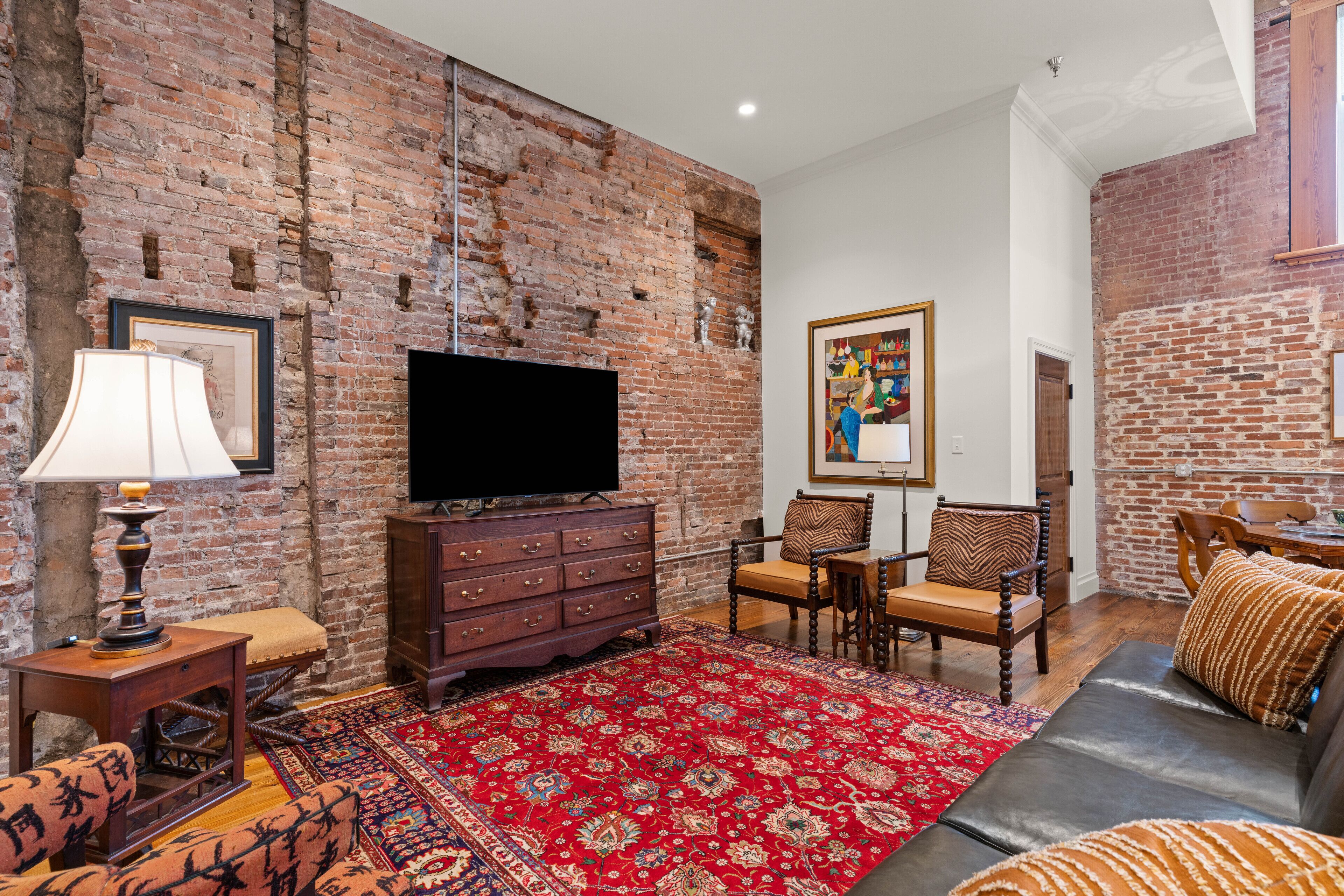 A living room with a brick wall, a flat-screen TV, wooden furniture, and a red patterned rug.