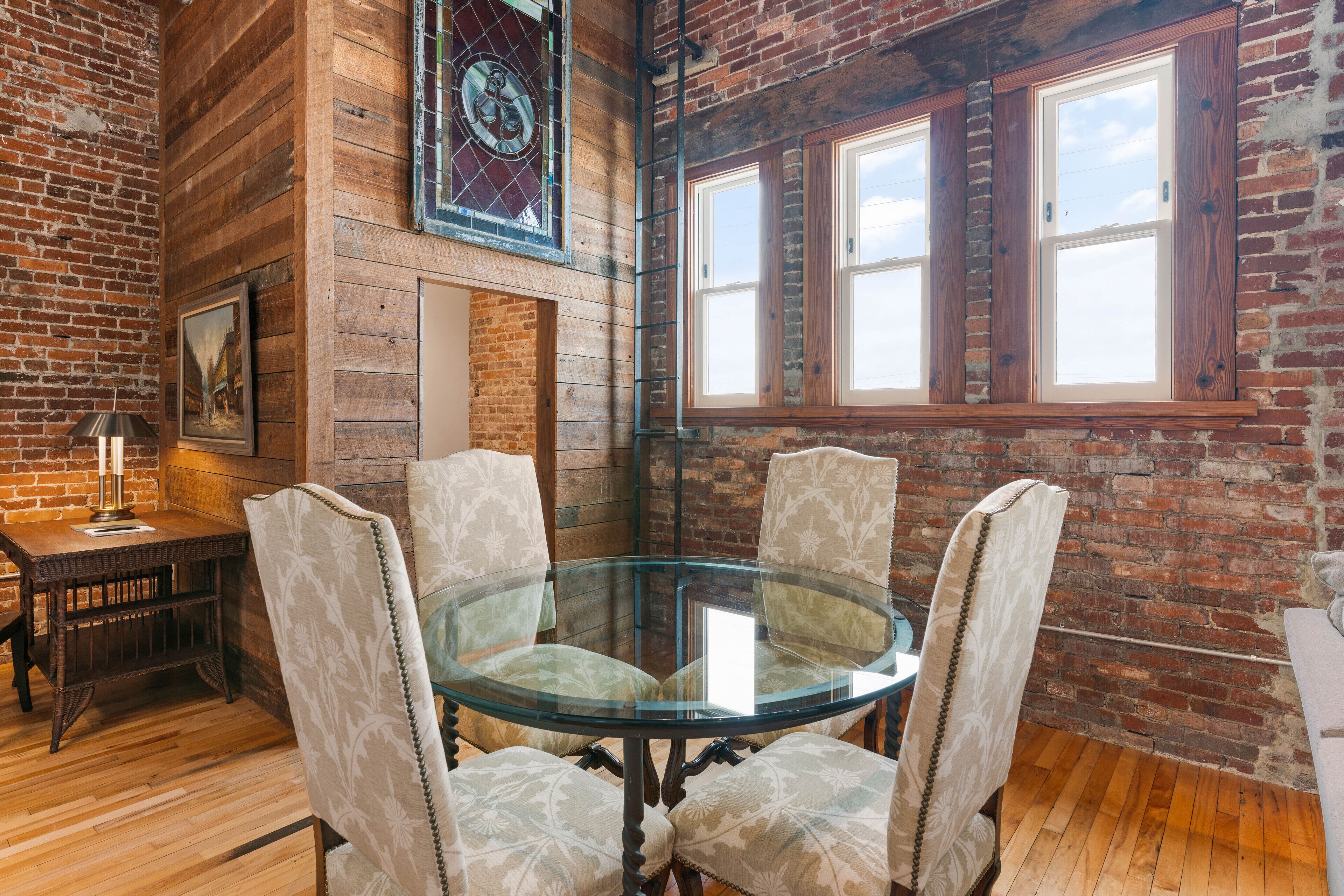 A round glass table with four upholstered chairs in a room with exposed brick walls and wooden floors.