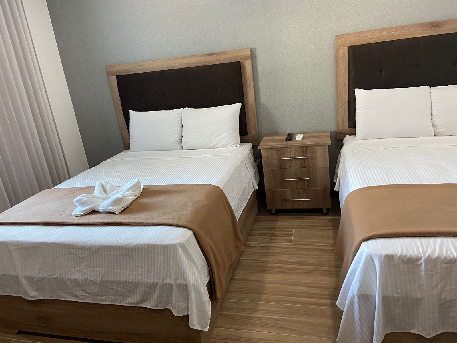 Quarto de hotel com duas camas, cada uma com cabeceira de madeira e roupa de cama branca. Existe uma mesinha de cabeceira de madeira entre as camas, com um controle remoto e um telefone.