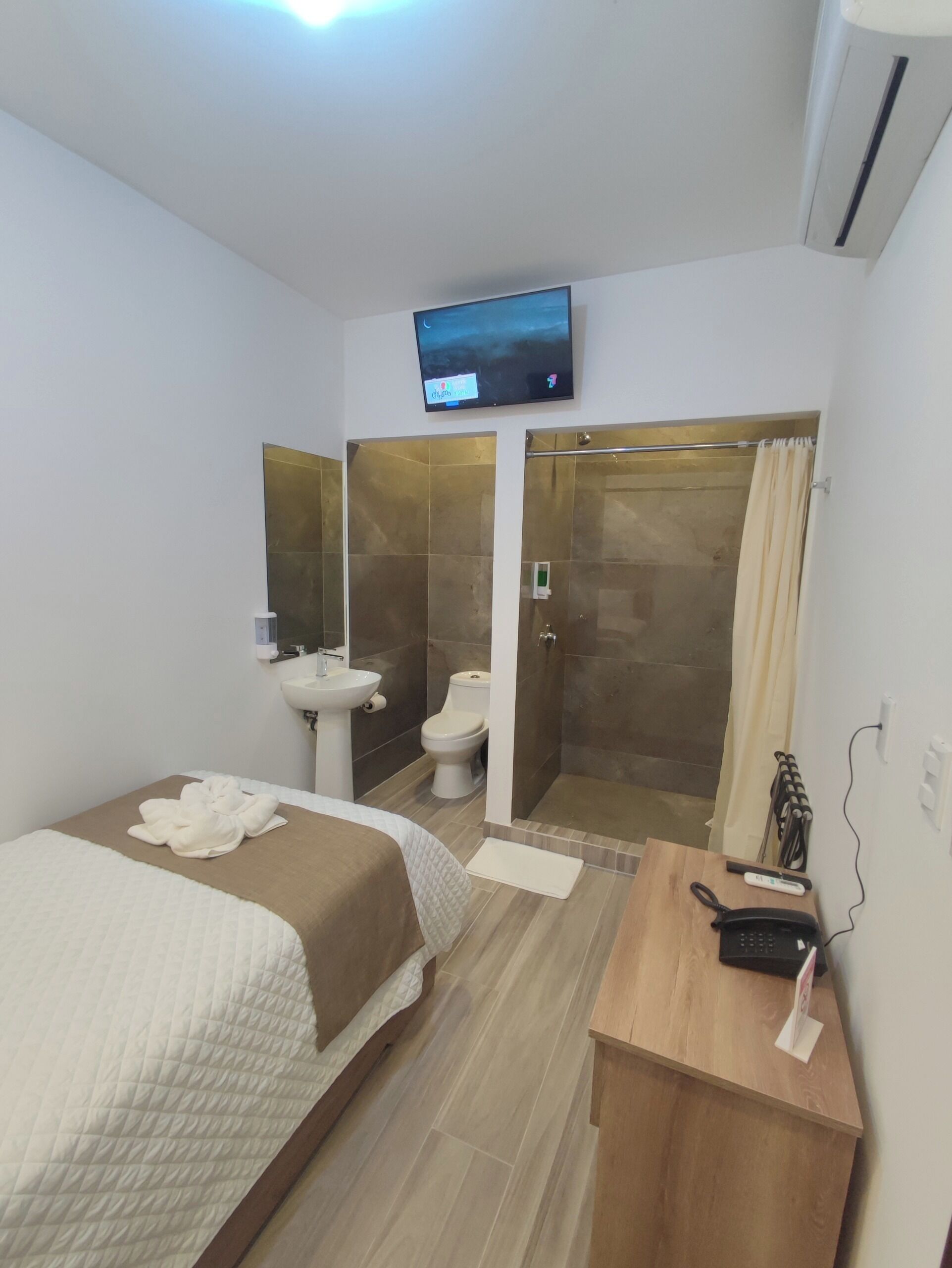 A hotel room with a bed, a bedside table with a phone, a TV mounted on the wall, a shower area, and a toilet.