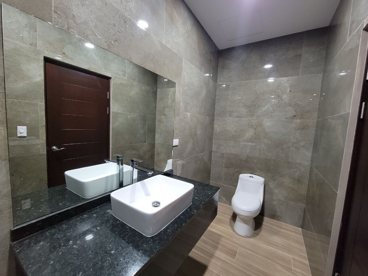 Un bagno moderno con uno specchio grande, due lavandini, un water e un piano di marmo scuro.