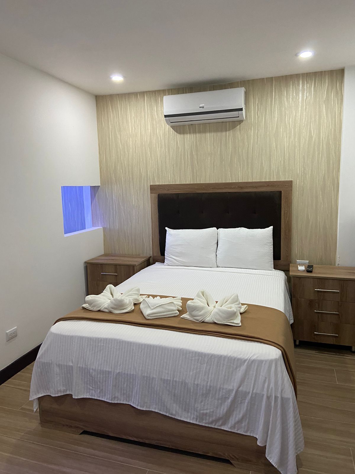 Una habitación de hotel con una cama, cabecera de madera, mesita de noche y equipo de aire acondicionado.