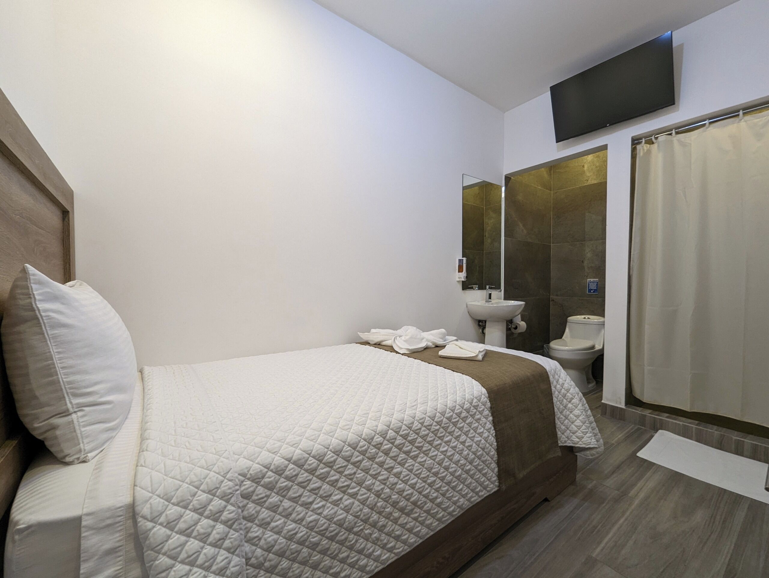 Una habitación de hotel moderna con cama, baño con lavamanos e inodoro, y un televisor de pantalla plana montado en la pared.