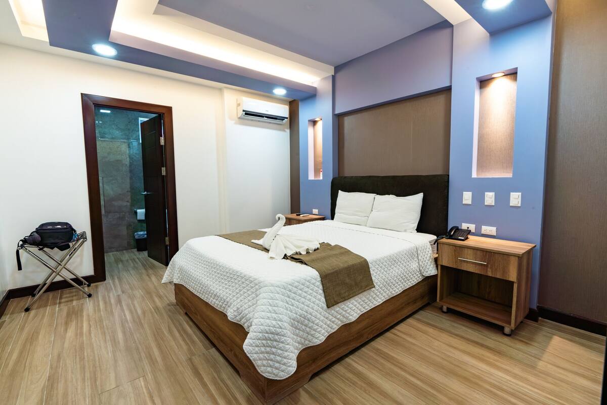Ein modernes Hotelzimmer mit einem großen Bett, Nachttischen, einem Kleiderschrank und einem durch eine geöffnete Tür einsehbaren Badezimmer.