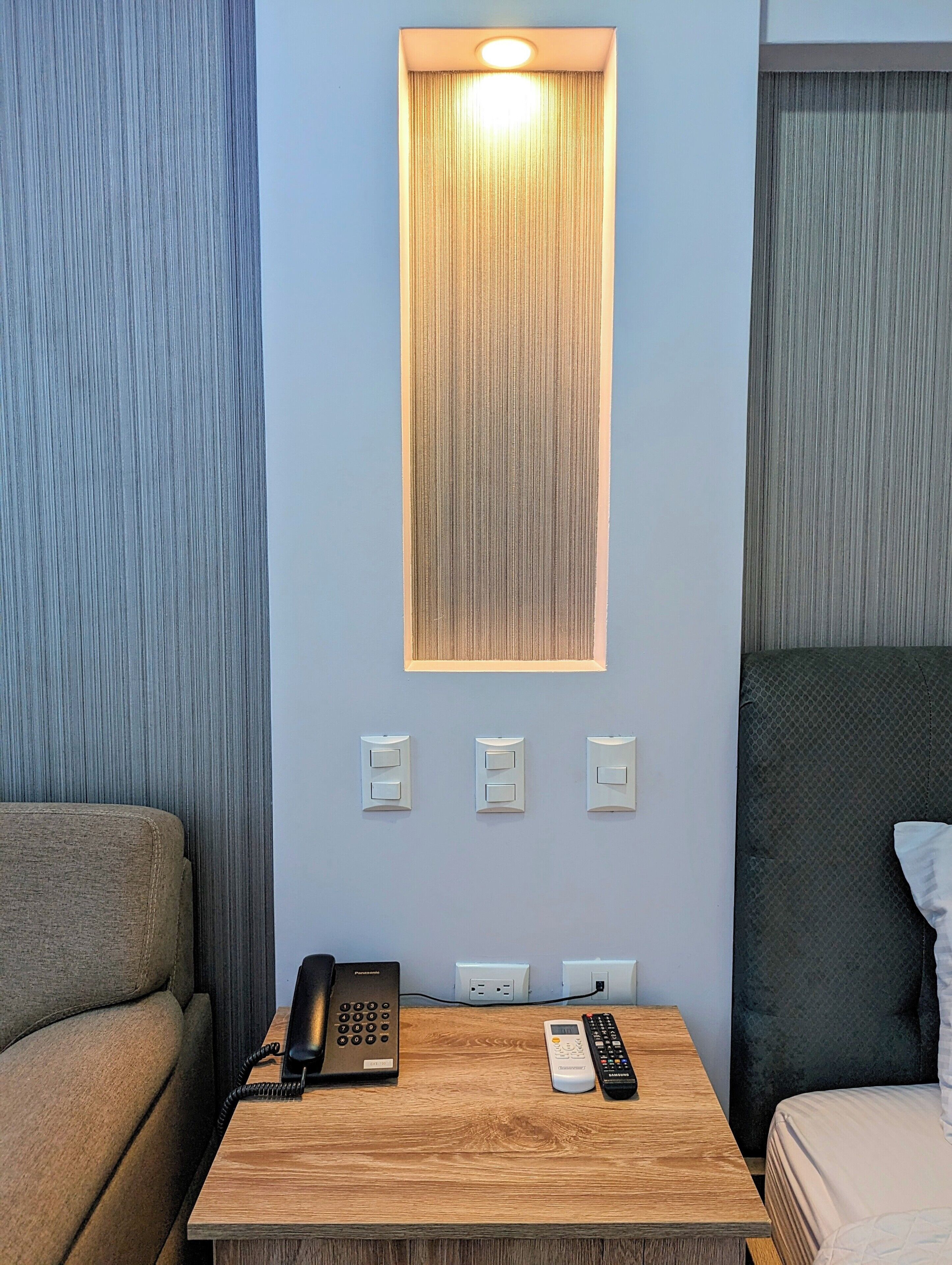 Habitación de hotel con mesita de noche de madera, teléfono, control remoto e iluminación empotrada en la pared.