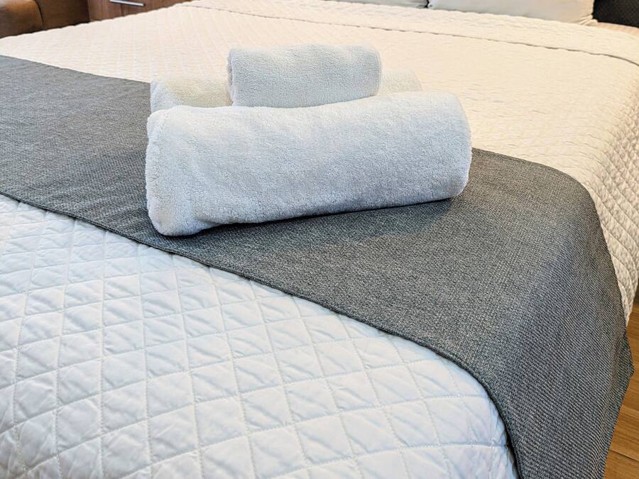 A neatly made bed with a quilted white duvet, a grey blanket, and two rolled white towels.