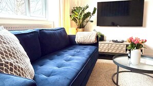 Un soggiorno con un divano blu, un tavolino rotondo, una televisione a parete e una finestra con tende.