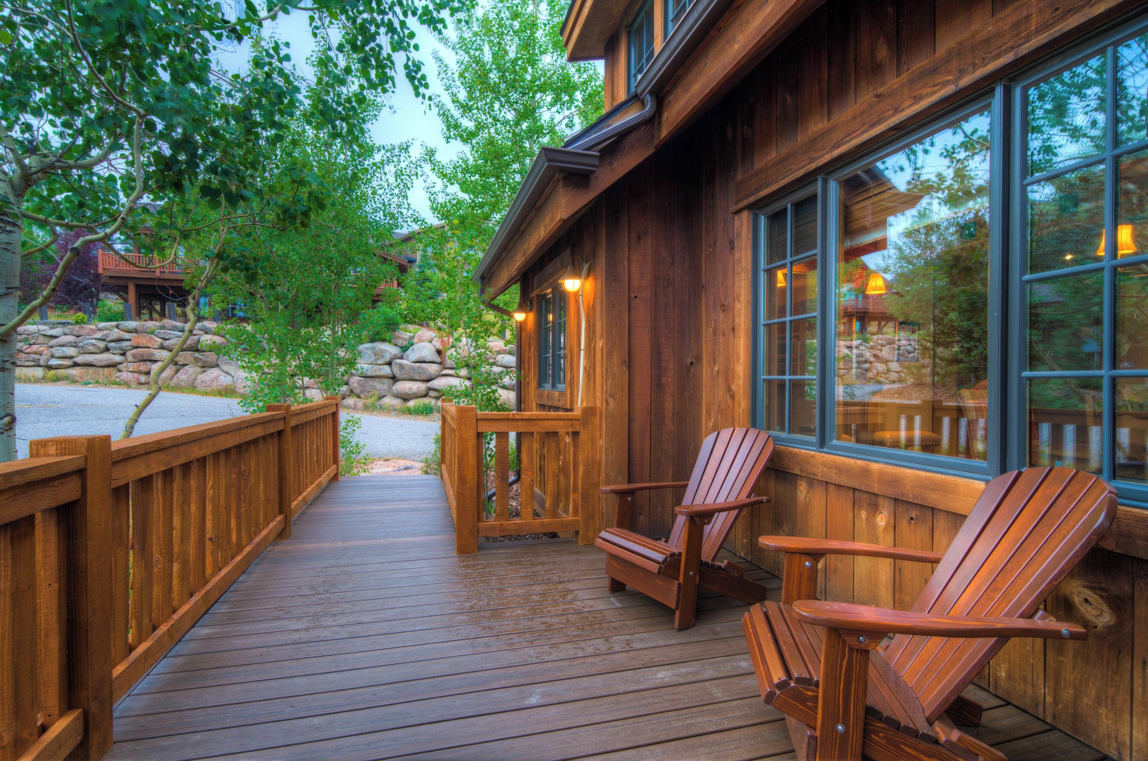 Una terraza de madera con sillas Adirondack, un muro de piedra y un edificio de madera con ventanas grandes.