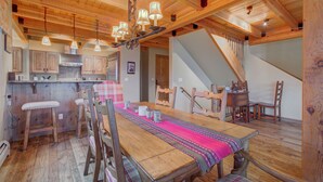 Ein Essbereich aus Holz mit einem langen Tisch, Stühlen und einer Chandelier. Im Hintergrund ist eine Küche mit Holzschränken und einer Hängelampe zu sehen.
