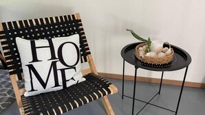 Ein Sessel mit einem 'HOME'-Kissen, ein kleiner runder Tisch mit einer Korb-Anordnung dekorativer Gegenstände und eine schlichte Wand.