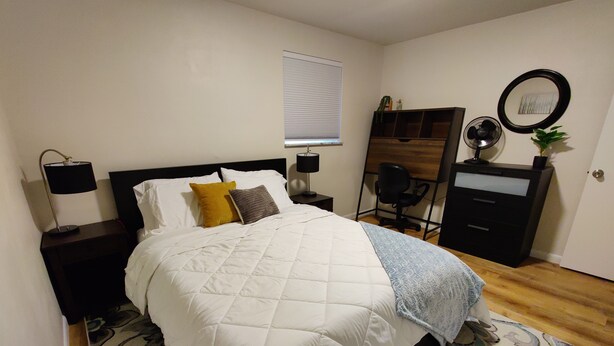 A neatly made bed with a quilt, a bedside lamp, a small desk with a lamp, a mirror, a dresser, and a chair.