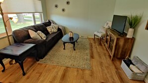 A living room with a dark brown sofa, a small round coffee table, a TV on a wooden stand, and a window with blinds.