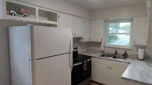 A compact kitchen with white cabinets, a refrigerator, a stove, and a window providing a view of the outside.