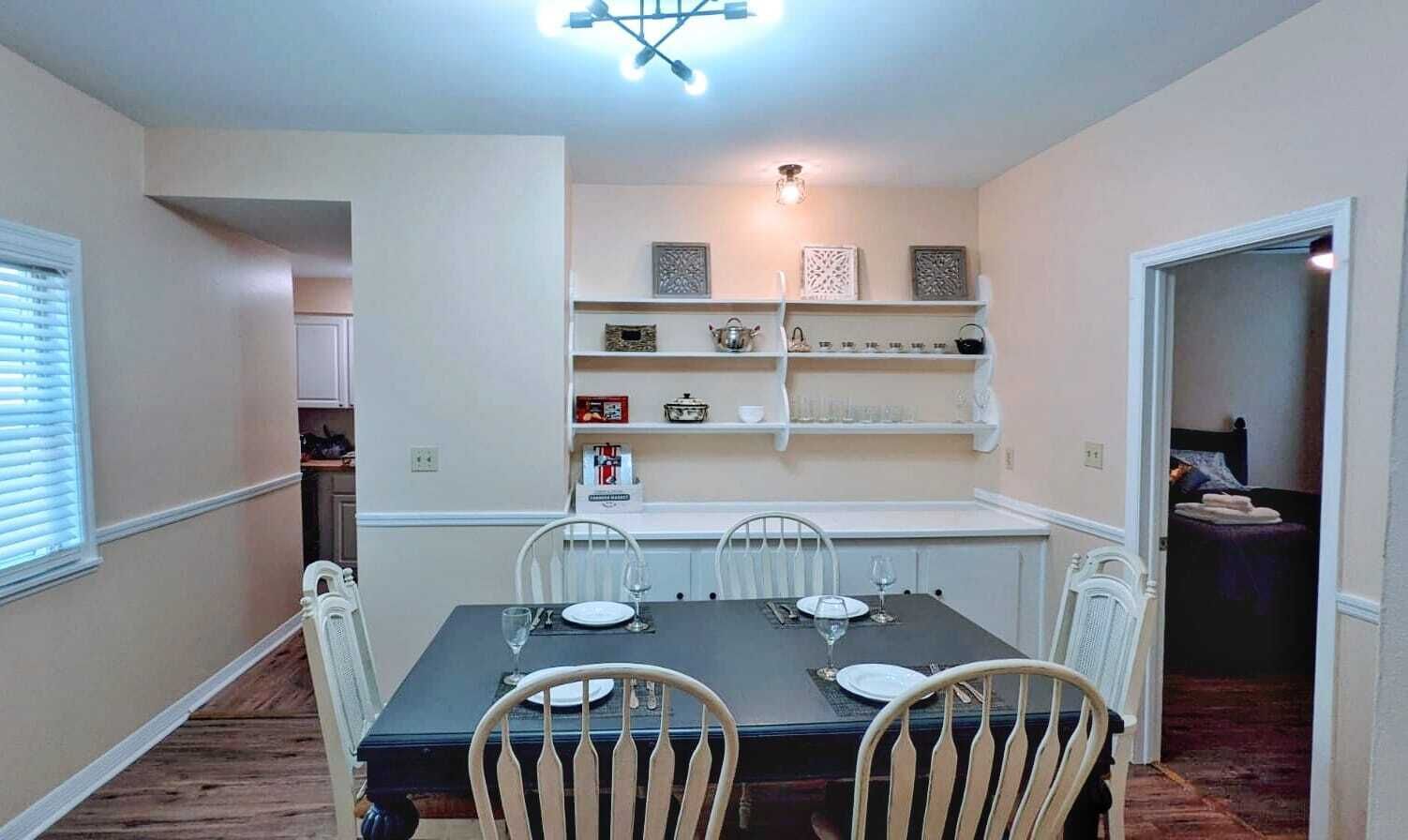 Una sala da pranzo con un tavolo rettangolare apparecchiato per quattro persone, circondato da sedie bianche. Sopra il tavolo, mensole con oggetti decorativi e una piccola sveglia.