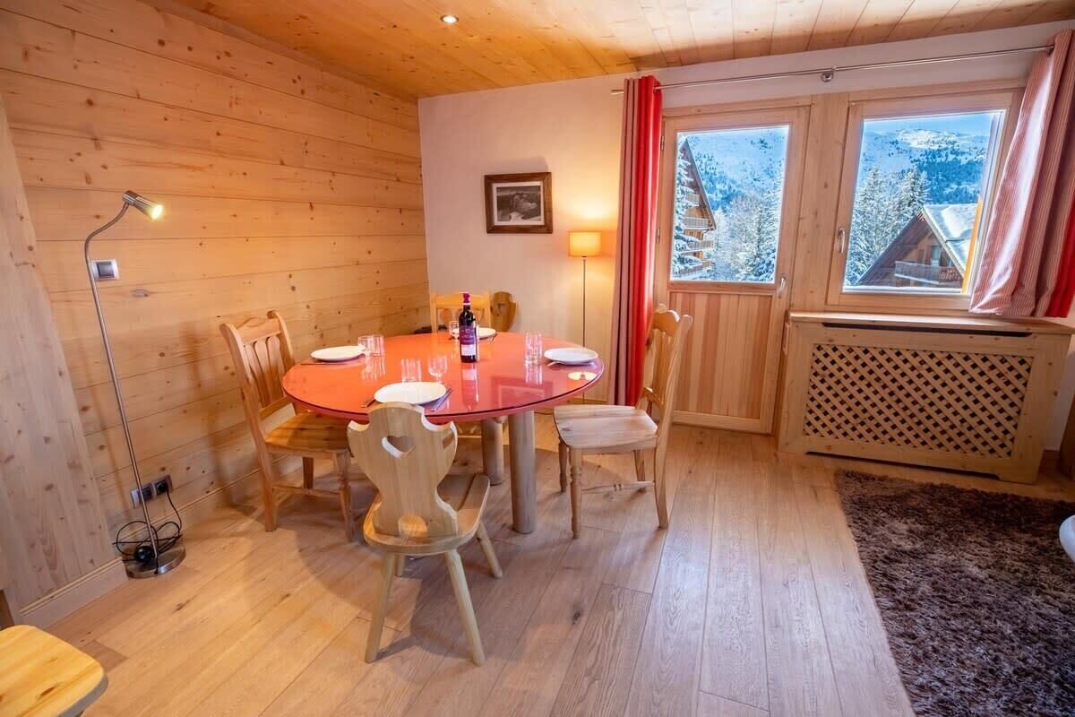 A wooden interior with a round table, chairs, a lamp, and a window with a view of snow-covered mountains.