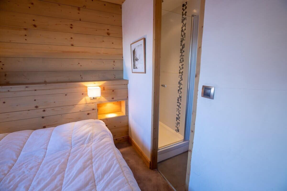 Une chambre avec un plafond en bois, un lit recouvert d’une couette blanche, une lampe de chevet et une cabine de douche en verre.