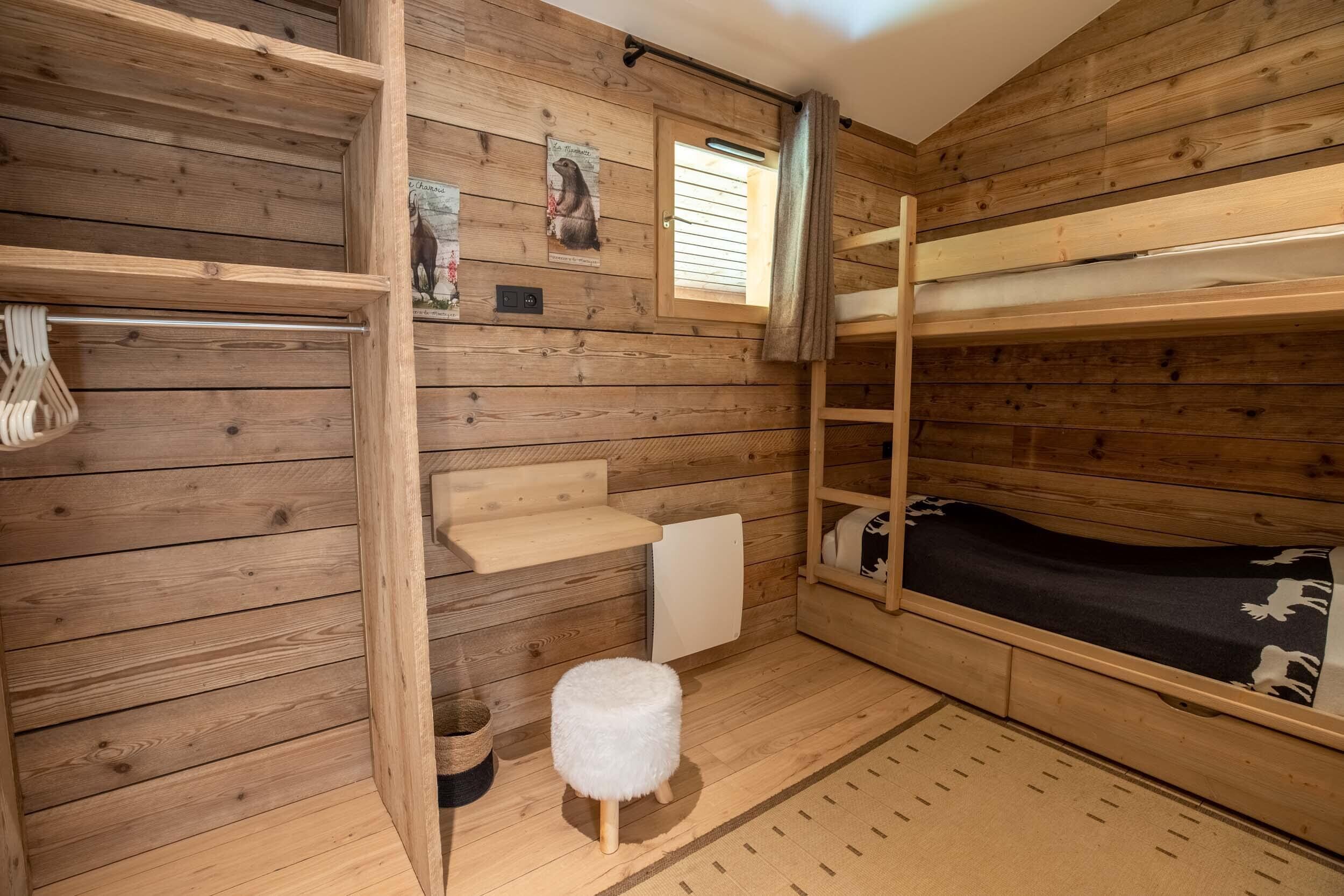 A wooden cabin room with bunk beds, a small stool, and a window with blinds.