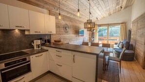 A modern kitchen with white cabinets, a built-in oven, and a coffee maker. There is a dining area with a sofa and chairs, and a large window with a view of the outdoors. - Sherpa 17 spacious apartment with sauna (Les Allues)