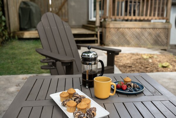 A patio with a table set for breakfast, including a coffee pot, a yellow mug, a plate of muffins, and a plate of fruit.