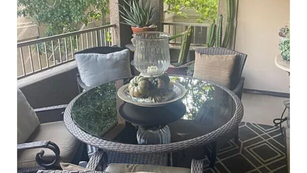 Uma mesa redonda com tampo de vidro, rodeada por cadeiras de vime e um banco estofado. Há um vaso de planta e um vaso decorativo com uma vela sobre a mesa.