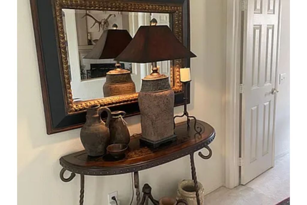 A hallway with a mirror, a table with decorative items, and a door.