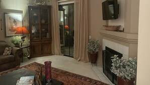 Uma sala de estar com lareira, televisão montada acima, sofá, mesa de centro com livros e um vaso com planta.