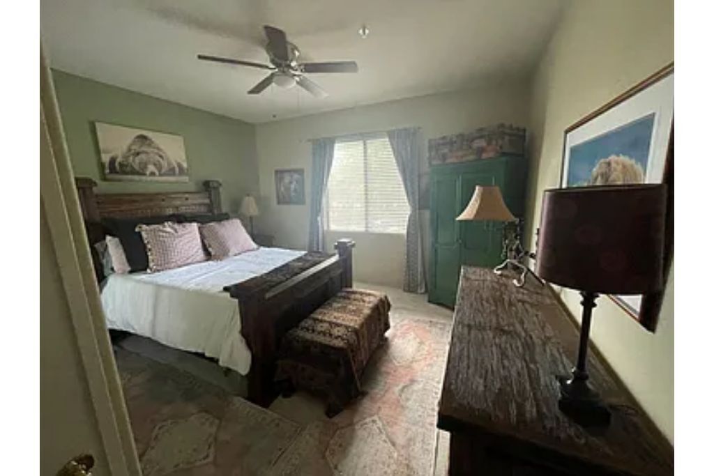 Une chambre à coucher comprenant un lit, un bureau, une chaise, un ventilateur de plafond, une fenêtre avec des rideaux et un tableau encadré accroché au mur.