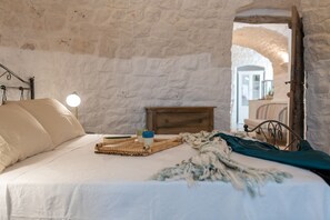 Un dormitorio con pared de piedra, una cama con ropa de cama blanca, una mesita de noche de madera y una lámpara.