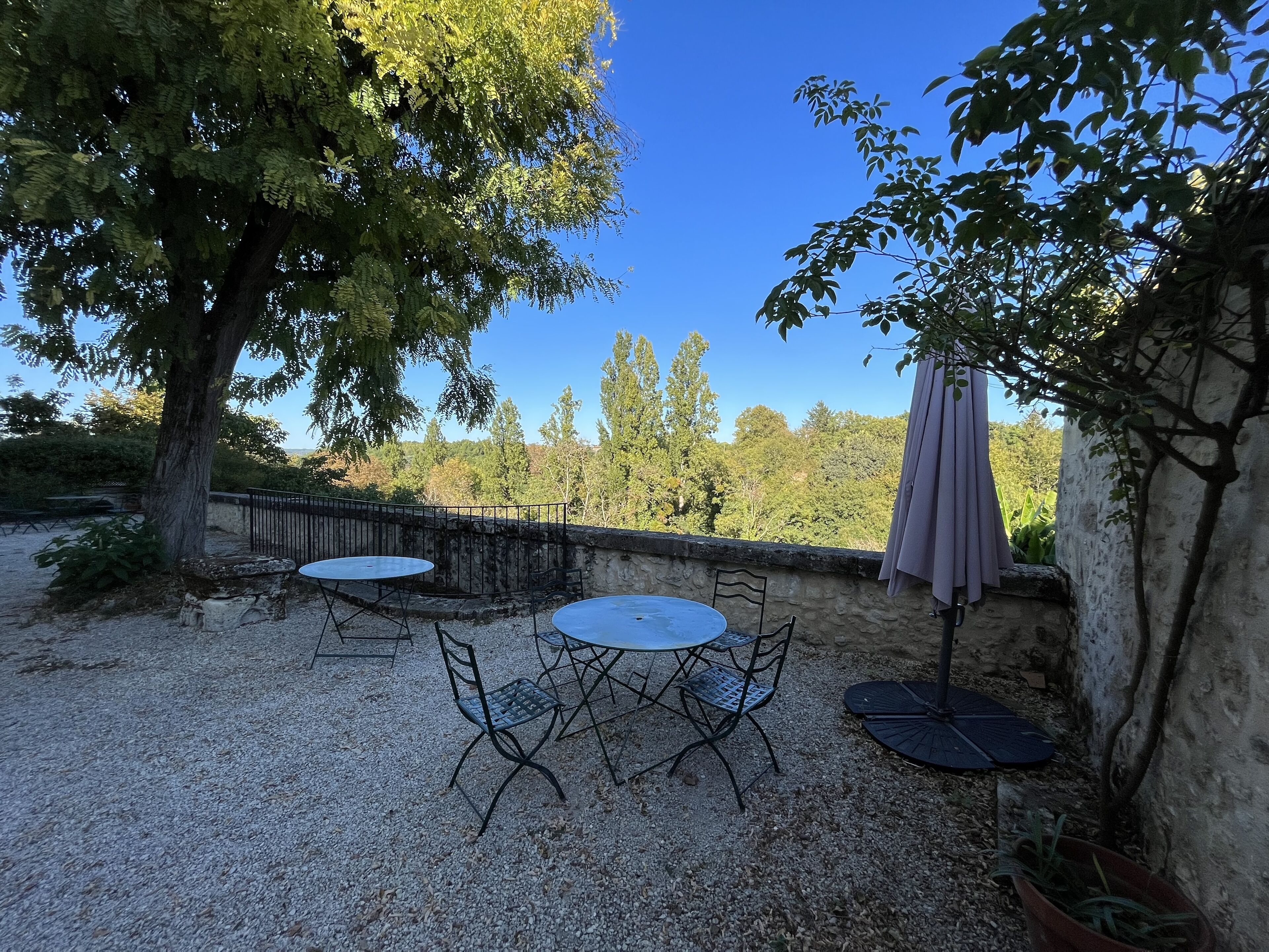 An outdoor patio with two round tables and chairs, surrounded by gravel, with a stone wall and a tree providing shade.